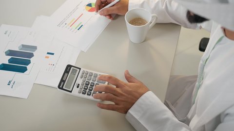 تحليل نتائج البيانات الاحصائية، تدوين الملاحظات على الورق، استخدام الالة الحاسبة لاجراء العمليات الحسابية، وظيفة مكتبية في شركات الامارات، احتساء القهوة في المكتب، تصوير علوي لرجل عربي خليجي اماراتي يرتدي الكندورة يجلس في مكتبه ويقوم بإنجاز مهام العمل