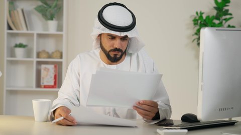 قراءة التقارير والنتائج، تطور الشركات الاماراتية، دمج العمل مع التقنية والتكنولوجيا، استخدام جهاز تقني حديث ومتطور،  إدارة شؤون الأعمال وتنظيمها، إتمام مهام العمل في المكتب، رجل عربي خليجي اماراتي يرتدي الكندورة والغترة يستخدم الحاسوب، مهنة ووظيفة مكتبية