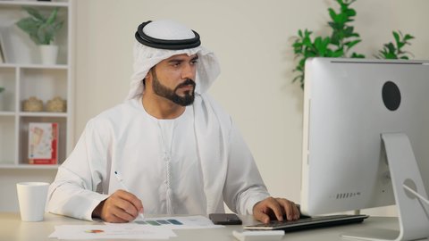 Integrating work with technology, using a modern and advanced technical device, taking notes on paper, managing and organizing business affairs, completing work tasks in the office, the development of Emirati companies, an Arab Gulf Emirati man wearing a kandura and ghutra using a computer, an office profession and job