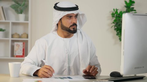 تطور الشركات الاماراتية، دمج العمل مع التقنية والتكنولوجيا، استخدام جهاز تقني حديث ومتطور، تدوين الملاحظات على الورق، إدارة شؤون الأعمال وتنظيمها، إتمام مهام العمل في المكتب، رجل عربي خليجي اماراتي يرتدي الكندورة والغترة يستخدم الحاسوب، مهنة ووظيفة مكتبية