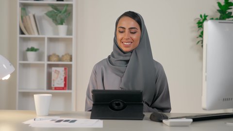 إجراء اجتماع عمل اونلاين، مهنة ووظيفة نسائية، ادارة الأعمال وانجاز المهام المطلوبة، تصوير مقرب لامرأة عربية خليجية اماراتية محجبة ترتدي عباءة ملونة تجلس خلف الطاولة تستخدم الجهاز اللوحي بإيماءات السرور، سيدة سعودية تستخدم جهاز تقني حديث ومتطور في المكتب