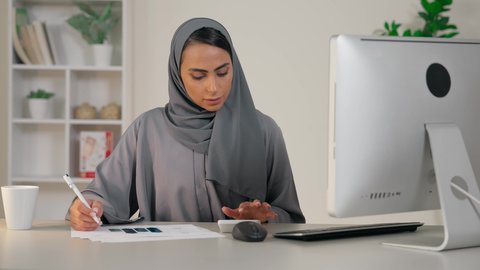 وظيفة مكتبية في شركات الخليج، تحليل نتائج البيانات الاحصائية، امراة عربية خليجية اماراتية محجبة ترتدي العباءة تجلس خلف الطاولة تقوم باستخدام الالة الحاسبة لاجراء العمليات الحسابية، انجاز المهام في المكتب، سيدة سعودية تدون الملاحظات على الورق