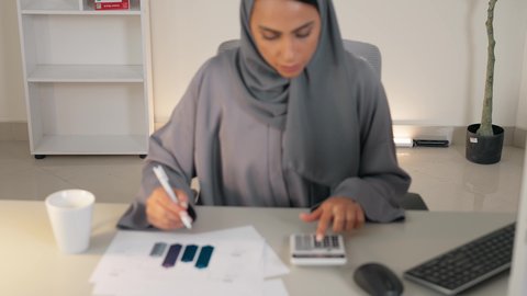 وظيفة مكتبية في شركات الخليج، تحليل نتائج البيانات الاحصائية، امراة عربية خليجية اماراتية محجبة ترتدي العباءة تجلس خلف الطاولة تقوم باستخدام الالة الحاسبة لاجراء العمليات الحسابية، انجاز المهام في المكتب، سيدة سعودية تدون الملاحظات على الورق