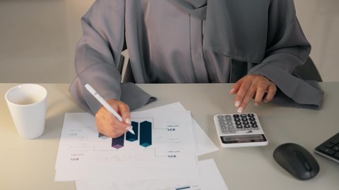 تحليل نتائج البيانات الاحصائية، تصوير مقرب لامراة عربية خليجية اماراتية محجبة ترتدي العباءة تجلس خلف الطاولة تقوم باستخدام الالة الحاسبة لاجراء العمليات الحسابية، انجاز المهام في المكتب، سيدة سعودية تدون الملاحظات على الورق، وظيفة مكتبية في شركات الخليج