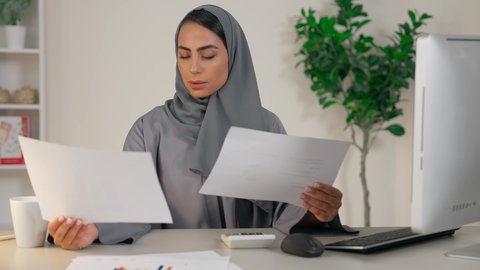 الاطلاع على المخططات، وظيفة مكتبية في شركات الخليج، تحليل نتائج البيانات الاحصائية، امراة عربية خليجية اماراتية محجبة ترتدي العباءة تجلس خلف الطاولة تقوم بمقارنة التقارير والنتائج، سيدة سعودية تقوم بإنجاز مهام العمل داخل المكتب
