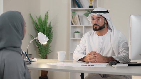 Emirati employee wearing kandura and ghutra sitting behind a desk, conducting a job interview, exchanging experiences and information to achieve goals, holding a meeting inside the company headquarters, understanding and cooperation between colleagues and developing strategic plans, two Arab Gulf Emirati employees learning work methods and professional development