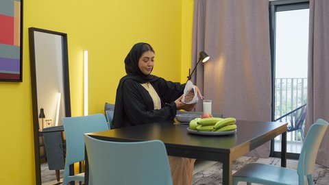 Preparing food that children love, preparing meals and fast food, gestures of happiness and pleasure, the joy of family gatherings at the table, a smiling veiled Emirati Gulf Arab woman wearing an abaya sitting at the table preparing a plate of cornflakes for her son