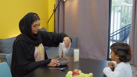 Spending quality time with the children, preparing meals and fast food, gestures of happiness and pleasure, the joy of the family gathering at the table, a smiling veiled Emirati Gulf Arab woman wearing an abaya sitting at the table preparing and arranging a plate of cornflakes for her son