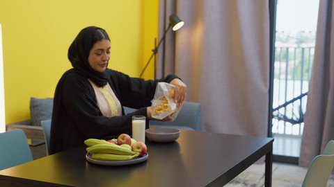 تجهيز الوجبات والمأكولات السريعة، ايماءات السعادة والسرور، متعة لمة الاهل على السفرة، امراه عربية خليجية اماراتية مبتسمة محجبة ترتدي العباءة تجلس على الطاوله بقوم بتحضير واعداد طبق الكورنفليكس لابنتها