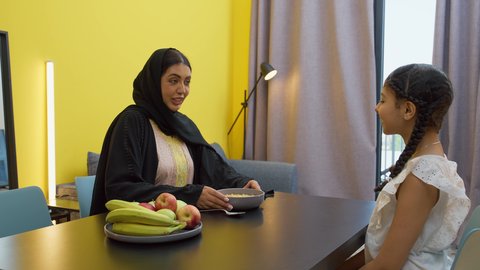 Gestures of happiness and pleasure, the joy of family gatherings at the table, a smiling veiled Emirati Gulf Arab woman wearing an abaya sitting at the table serving a plate of cornflakes to her daughter, a Gulf girl eating a healthy snack