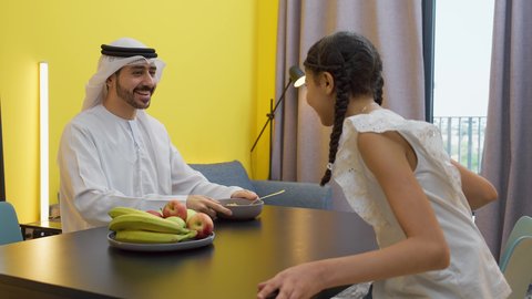مفهوم الأبوة والاهتمام بالأولاد، تناول وجبة صحية، إفطار صباحي عائلي، رجل عربي خليجي اماراتي يرتدي الكندورة و الغتره  يجلس على الطاوله يعطي وجبه سناك صحيه لابنته، العطف والحنان بين أفراد الأسرة، طفلة خليجيه تقوم بتناول سناكس