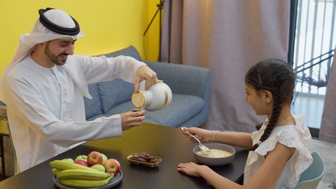 اجواء عائلية سعيدة، تناول وجبة صحية، إفطار صباحي عائلي،  أب عربي خليجي إماراتي يجلس على الطاولة يقوم بصب كوب من القهوه برفقه ابنته، العطف والحنان بين أفراد الأسرة، طفلة خليجيه تقوم بتناول الكورن فليكس بالحليب