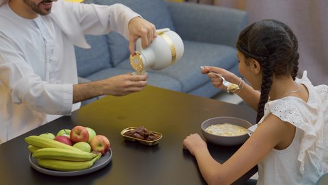 Affection and tenderness between family members, a happy family atmosphere, eating a healthy meal, a family breakfast, an Emirati Gulf Arab father sitting at the table pouring a cup of coffee with his daughter, a Gulf girl eating cornflakes with milk