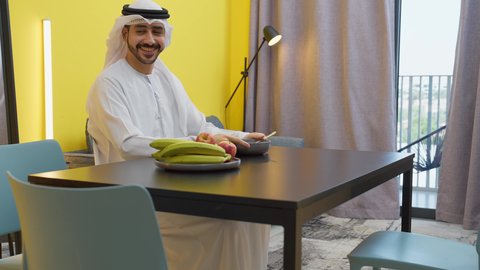 Eating a healthy meal, fresh healthy fruits, meals and food, an Emirati Gulf Arab father wearing a kandura and ghutra sitting at the table inside the living room with his son offering a plate of chocolate beans to his son