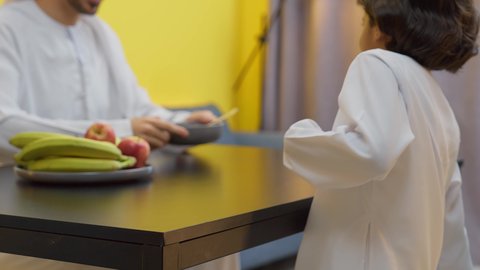 تناول وجبة صحية، الفواكه الصحية الطازجة، وجبات ومأكولات غذائية، اب عربي خليجي اماراتي يرتدي الكندورة و الغتره يجلس على الطاوله داخل عرفة المعيشة برفقه ابنه يقوم بتقديم طبق من حبوب الشوكولاته لابنه
