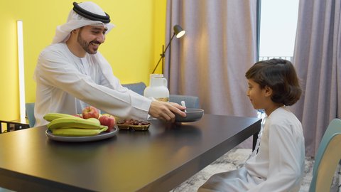 Healthy meals and foods, fresh healthy fruits, an Emirati Gulf Arab father wearing a kandura and ghutra sitting at a table inside the living room with his son pouring coffee with gestures of happiness and pleasure, a Gulf child eating a plate of chocolate beans