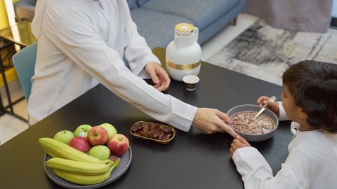الفواكه الصحية الطازجة، وجبات ومأكولات صحية، اب عربي خليجي اماراتي يرتدي الكندورة و الغتره يجلس على الطاوله داخل عرفة المعيشة برفقه ابنه يقوم بصب القهوة بايماءات السعاده و السرور، تصوير مقرب لطفل خليجي يقوم بتناول طبق من حبوب الشوكولاته