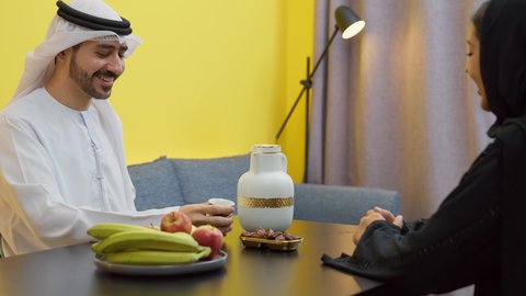 The concept of affection and love between spouses, mood adjustment by consuming caffeine, the concept of family bonding and understanding, an Emirati Gulf Arab man wearing the Emirati kandura and ghutra sitting at the table in the living room with his wife, pouring a cup of Arabic coffee with expressions of happiness and pleasure