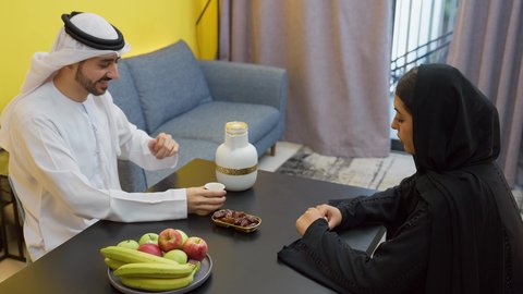 Affection and love between spouses, mood adjustment by consuming caffeine, the concept of family bonding and understanding, an Arab Gulf Emirati man wearing the Emirati kandura and ghutra sitting at the table in the living room with his wife pouring a cup of Arabic coffee with expressions of happiness and pleasure