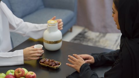 الاستمتاع بتناول الكافيين، المودة والمحبة بين الزوجين، مفهوم الترابط و التفاهم الأسري، رجل عربي خليجي اماراتي يرتدي الكندورة والغترة الإماراتية يجلس على الطاوله فى غرفه المعيشة برفقه زوجته يقوم بصب  فنجان من القهوه العربية بتعابير السعادة والسرور