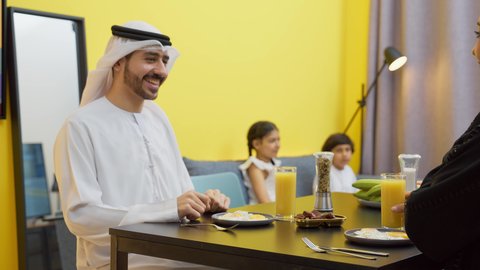 Having a good time, strengthening the bonds of love and family cohesion, an Arab Gulf Emirati family sitting in the living room inside the house, an Arab Gulf Emirati man wearing a kandura and ghutra sitting at the dining table with his wife eating breakfast
