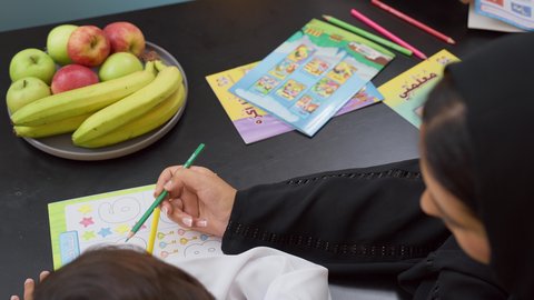 مفهوم الأمومة والطفولة، مفهوم فنون الرسم عند الأطفال ، قضاء اوقات عائلية ممتعة في المنزل،  ام عربيه خليجيه اماراتية ترتدي العباءة السوداء تجلس على الطاوله فى غرفه المعيشة تقوم بتعليم ومشاركة ابنها بالتلوين،تعلم الفن و التطوير والإبداع