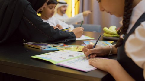 عائله عربيه خليجيه اماراتية ترتدي الزي التقليدي يجلسون في غرفه المعيشه مع ابنائهم بأيماءات السعادة و السرور، التعلم والابداع وتنمية المهارات، قضاء اوقات عائلية ممتعة في المنزل، تصوير مقرب لفتاة عربيه خليجيه تقوم باداء واجباتها المدرسية