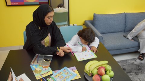 مفهوم الأمومة والطفولة، مفهوم فنون الرسم عند الأطفال ، قضاء اوقات عائلية ممتعة في المنزل،  ام عربيه خليجيه اماراتية ترتدي العباءة السوداء تجلس على الطاوله فى غرفه المعيشة تقوم بتعليم ومشاركة ابنها بالتلوين،تعلم الفن و التطوير والإبداع