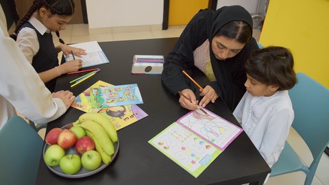 الأنشطة التعليمية وتنميه قدرات الابناء بالتعلم، الاهتمام بتعليم الأبناء، دعم وتشجيع الاطفال لحل الدروس والواجبات المدرسيه المنزليه،عائله عربيه خليجيه اماراتية ترتدي الزي التقليدي تجلس على الطاوله وتساعد ابنائها  في التعليم والقراءه