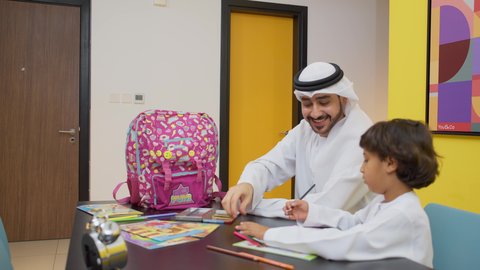 البهجة بالعودة إلى المدرسة, عائله عربيه خليجيه اماراتية تقوم بالاستعداد و التحضير لبداية عام دراسي جديد، ام خليجيه ترتدي العباءه السوداء تقوم بوضع الكتب و الدفاتر داخل الحقيبة المدرسية برفقه ابنتها, اب عربي خليجي يرتدي الكندورة و الغتره يجلس مع ابنه على الطاوله