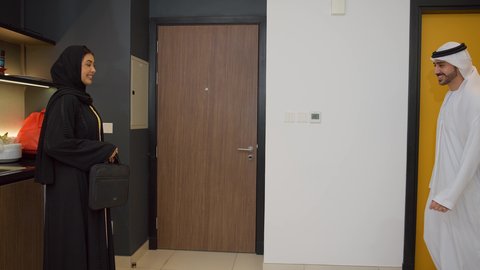 الاستعداد للذهاب إلى دوام العمل بنشاط, رجل عربي خليجي اماراتي يرتدي الكندورة والغترة يأخد حقيبه العمل من زوجته, الاهتمام بالأناقة والمظهر الخارجي، سيده اماراتية تقوم بمساعدة زوجها اثناء ذهابه إلى العمل بإيماءات السرور والسعادة
