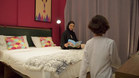 Teaching children to be educated and read, a Saudi woman spending quality time with her children, a passion for reading books and novels, doing fun activities at home, an Emirati Gulf Arab mother wearing a black abaya sitting on the bed next to her son and reading stories and tales before bed