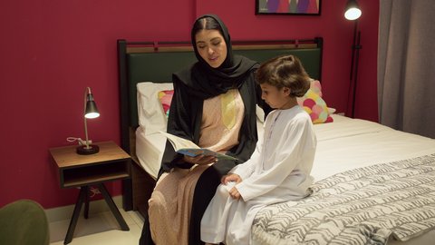 شغف المطالعة للكتب والروايات، القيام بالانشطة المسلية في المنزل، تعليم الاولاد على التثقيف والمطالعة، سيده سعودية تقوم بقضاء وقت ممتع مع الأبناء، ام عربية خليجيه اماراتية ترتدي العباء السوداء تجلس على السرير بجانب ابنها و تقوم بقراءة القصص والحكايات قبل النوم
