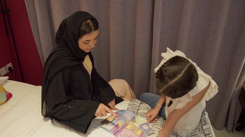 مفهوم التربيه القائمه على الحب والحنان، تعزيز الروابط العائلية، ام سعودية تقوم بقضاء وقت ممتع مع الابنه، المشاركه بقراءه القصص المفيده مع الأبناء قبل النوم، تصوير علوي لامراة عربيه خليجيه اماراتية ترتدي العباءه تجلس على السرير برفقه ابنتها تقرأ الروايات المسليه