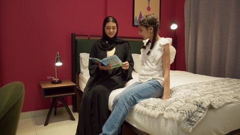 ام سعودية تقوم بقضاء وقت ممتع مع الابنه، المشاركه بقراءه القصص المفيده مع الأبناء قبل النوم، مفهوم التربيه القائمه على الحب والحنان، تعزيز الروابط العائلية، امراة عربيه خليجيه اماراتية ترتدي العباءه تجلس على السرير برفقه ابنتها تقرأ الروايات المسليه
