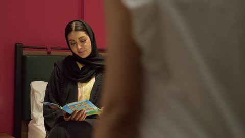 تعزيز الروابط العائلية، مفهوم التربيه القائمه على الحب والحنان، ام سعودية تقوم بقضاء وقت ممتع مع الابنه، المشاركه بقراءه القصص المفيده مع الأبناء قبل النوم، بامراة عربيه خليجيه اماراتية ترتدي العباءه السوداء تجلس على السرير برفقه ابنتها تقرأ الروايات المسليه