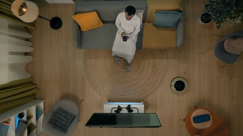 اضاءة زرقاء ثلاثية الابعاد ، بورتريه علوي عامودي لشاب عربي خليجي سعودي يرتدي الثوب والشماغ التقليدي يجلس على الاريكة يحمل بيده الهاتف المحمول، ربط الادوات التقنية ببعضها باستخدام الجوال