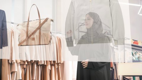 متعة الشراء في المولات السعودية، اختيار الملابس والتسوق بالتقنية والتكنولوجيا المستقبلية الحديثة, امرأة عربية خليجية سعودية محجبة ترتدي عباءة سوداء تقوم بالتفاعل مع شاشة هولوجرامية ثلاثية الابعاد لعرض الألبسة، رؤية مستقبيلة سعودية 2032 ، خلفية متجر ملابس