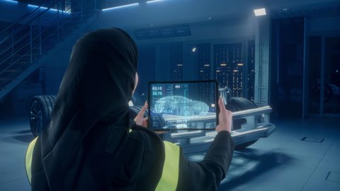 مفهوم الهندسة الصناعية، العمل في شركات التقنية والتكنولوجيا المستقبلية الحديثة, تصوير من الخلف لمهندسة عربية خليجية سعودية ترتدي سترة الحماية تقوم باستخدام جهاز التابليت المحمول، سيارة هولوجرامية ثلاثية الأبعاد، رؤية مستقبيلة سعودية 2030، خلفية مقر العمل