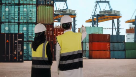 Port of loading goods and commercial containers, supervising the delivery and transportation of containers, using modern machines in import and export, future vision 2030, a rear view of a Saudi Arabian Gulf engineer wearing a safety helmet standing in the workplace with an engineer, the concept of monitoring the progress of projects