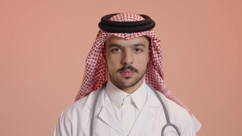 Concept of medicine and healthcare, portrait of a smiling Saudi Arabian Gulf Arab doctor wearing traditional thobe and shemagh with white coat, wearing a stethoscope, looking at the camera with expressions of happiness and pleasure, standing straight with crossed hands gestures
