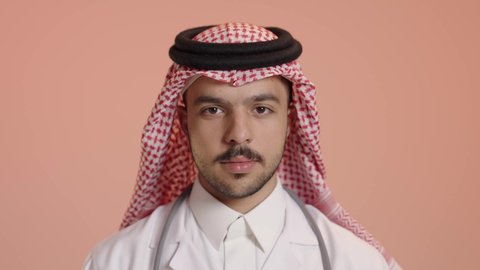 Readiness to provide services to patients with all efficiency, the concept of medicine and healthcare, portrait of a smiling Saudi Arabian Gulf Arab doctor wearing the traditional thobe and shemagh with a white coat, wearing a stethoscope, looking at the camera with expressions of happiness and pleasure, standing straight with crossed hands gestures