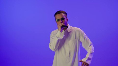تسجيل مقطع صوتي غنائي, الاستمتاع بأداء المواهب الصوتية، بورتريه لرجل عربي خليجي سعودي سعيد يرتدي الزي التقليدي و نظارة شمسية، يحمل الميكروفون و يغني بحماس، ايماءات وجه و يدين تدل على السعادة و الاستمتاع, ممارسة موهبة الغناء