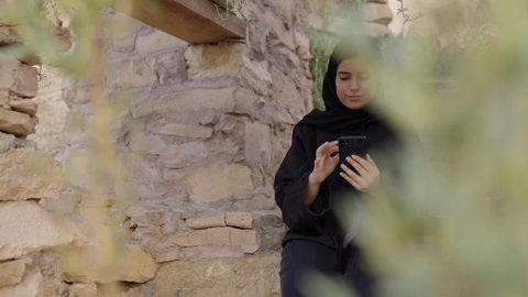 تصفح المواقع الالكترونية والتواصل الاجتماعي،  قضاء وقت ممتع فى الخارج, تصويرلامراة عربيه خليجيه سعودية ترتدي الحجاب و العباءة السوداء تجلس فى الهواء الطلق تستخدم الهاتف المحمول ، استخدام البرامج والتطبيقات الالكترونية، التسوق الالكتروني عبر الانترنت