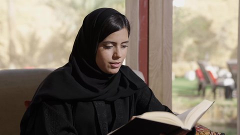 Culture, knowledge, and self-development, cultural recreational activities, the passion for engaging in beneficial hobbies, a depiction of a Gulf Arab Saudi woman wearing an abaya and hijab sitting on the sofa in the living room reading and studying.