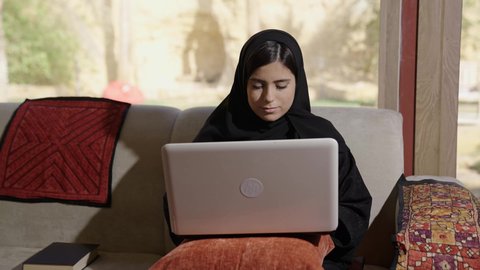 Gestures of integration and focus, content creation, the concept of data entry, a depiction of a Gulf Arab Saudi woman sitting in the living room using a laptop, typing on the laptop, integrating modern technology into practical life.