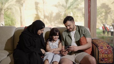 مفهوم الدفء والحب عائلي، استخدام الهاتف المحمول وتصفح المواقع الالكترونية، عائله عربيه خليجيه سعودية تجلس على الاريكه داخل غرفه المعيشه يتشاركون مشاهدة الهاتف بايماءات السعادة و السرور