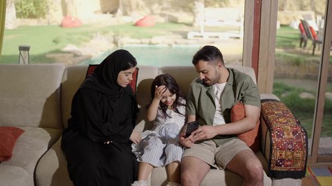 A warm and bonding family atmosphere, a Saudi Gulf Arab family sitting on the couch with their little daughter, laughing and raising her hands in gestures of happiness and joy, embodying the concept of affection and family delight.