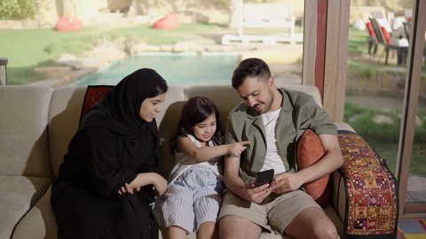 أجواء ترابط وأسري دافئ، استخدام الهاتف المحمول وتصفح المواقع الالكترونية، عائله عربيه خليجيه سعودية تجلس على الاريكه داخل غرفه المعيشه يتشاركون مشاهدة الهاتف بايماءات السعادة و السرور