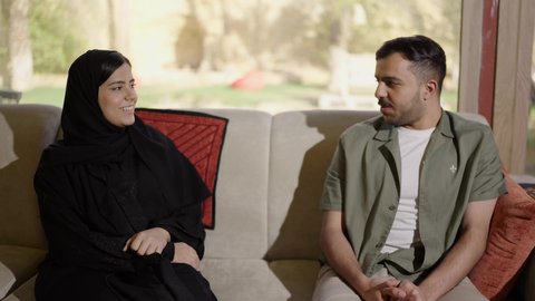 مفهوم التواصل العائلي، رجل عربي خليجي سعودي يرتدي الزي الكاجوال يجلس على الاريكه بجانب زوجته يتبادلان الحوار، جلسة عائلية دافئة، ايماءات السعاده و السرور